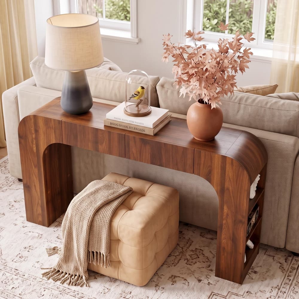 A modern curved wooden console table in a living room setting, styled with a table lamp, books, a small bird figurine under a glass dome, a vase of dried pink leaves, and a tufted ottoman draped with a beige throw blanket.