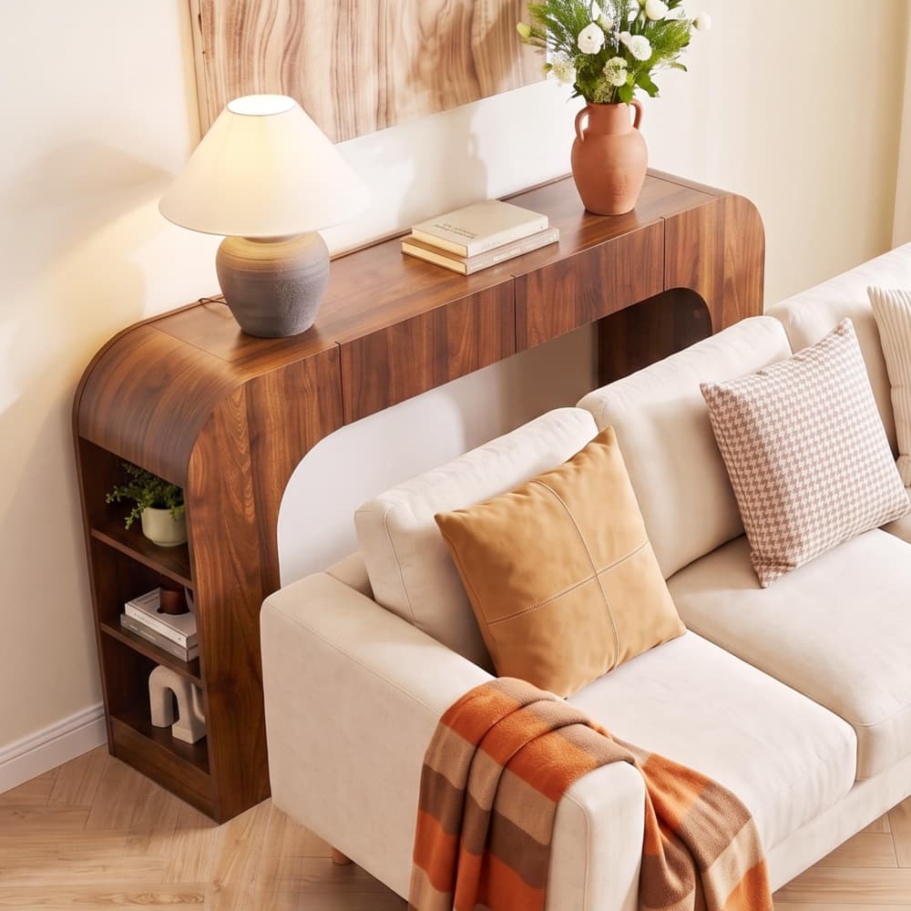 A modern curved wooden console table styled in a living room next to a cream sofa, holding a lit table lamp, books, and a terracotta vase with flowers.