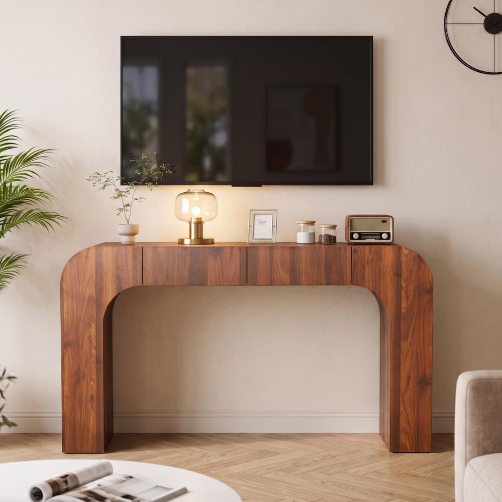 A modern curved wooden console table with a warm walnut finish, styled in a living room setting under a wall-mounted TV, with decorative items on top.