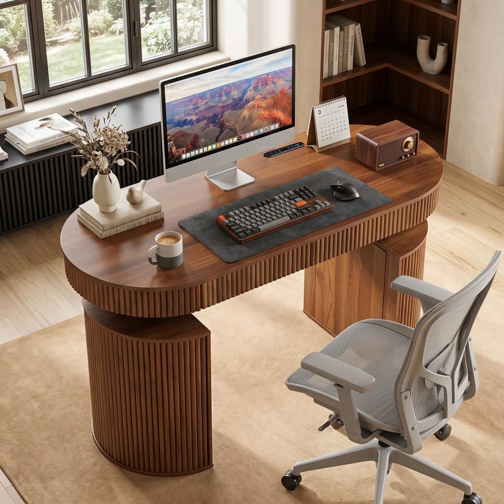 A modern oval wooden desk with fluted pedestal legs in a warm medium brown finish, styled in a bright home office with a gray mesh office chair, computer monitor, keyboard, and other desk accessories.