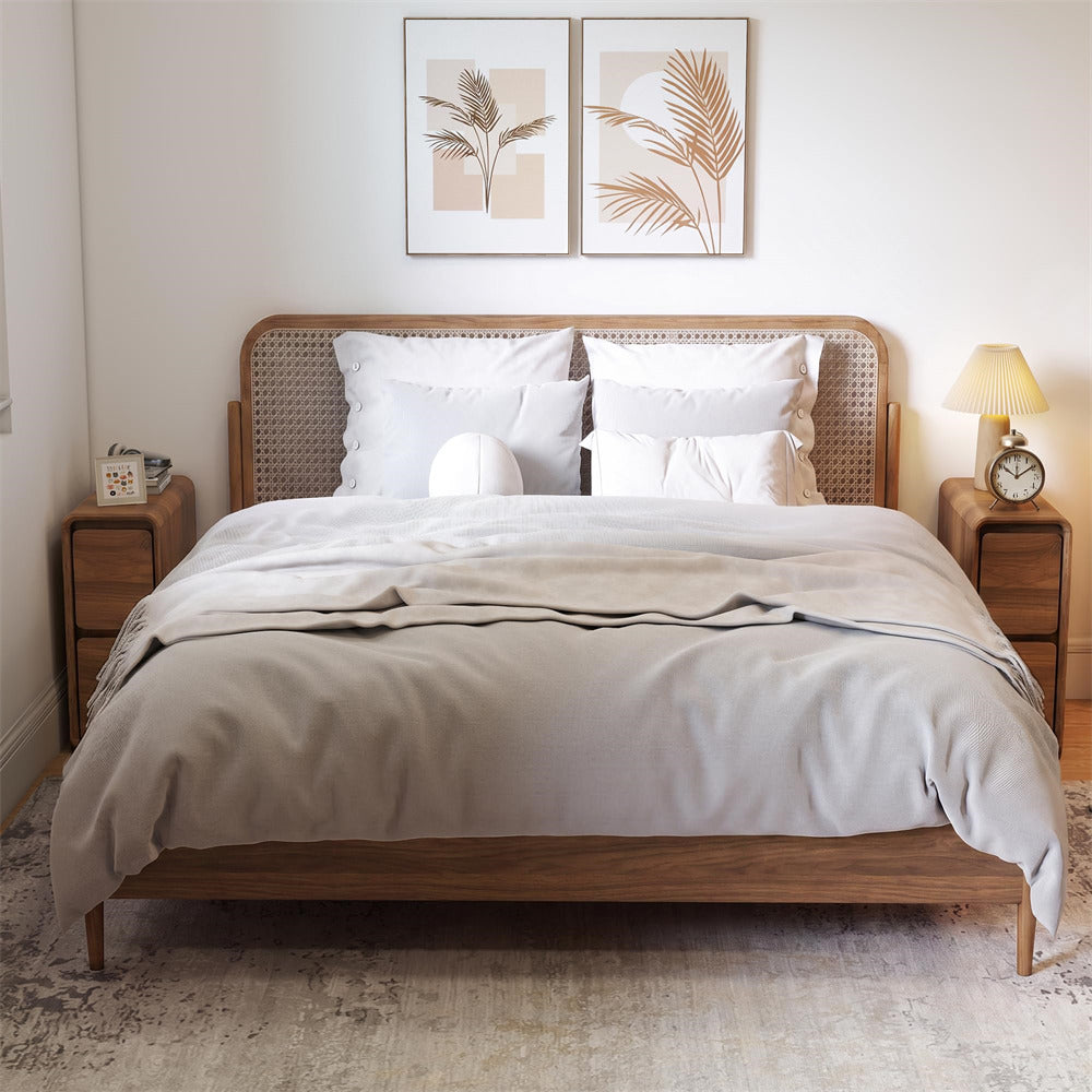 A neatly made bed with white linens and a light beige duvet, set in a bright bedroom with a wooden cane and wood frame bed, matching nightstands, a lamp, and two framed botanical prints on the wall.