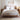 A neatly made bed with white linens and a light beige duvet, set in a bright bedroom with a wooden cane and wood frame bed, matching nightstands, a lamp, and two framed botanical prints on the wall.