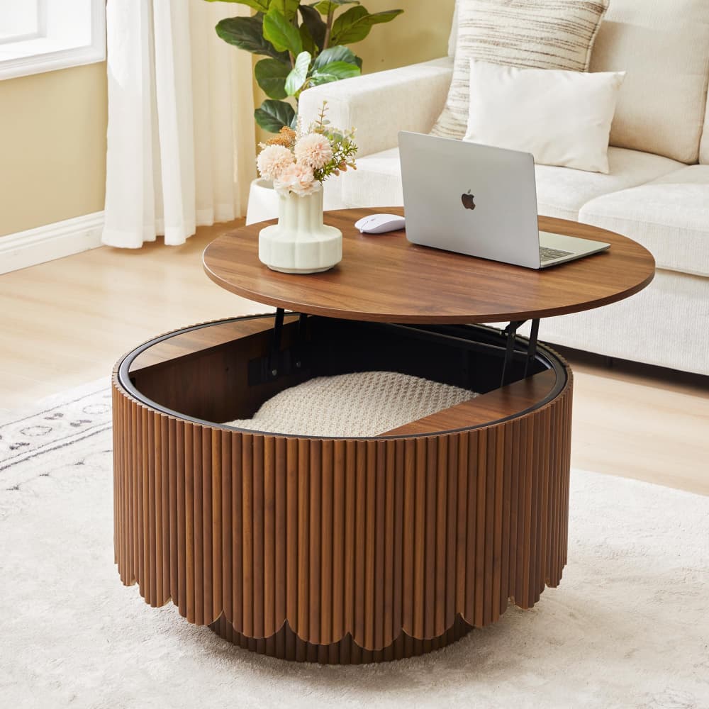 A modern round coffee table in a warm medium brown wood finish, featuring a fluted slatted cylindrical base with a built-in cream cushion and a hinged top surface holding a laptop and vase of flowers, set in a bright living room.