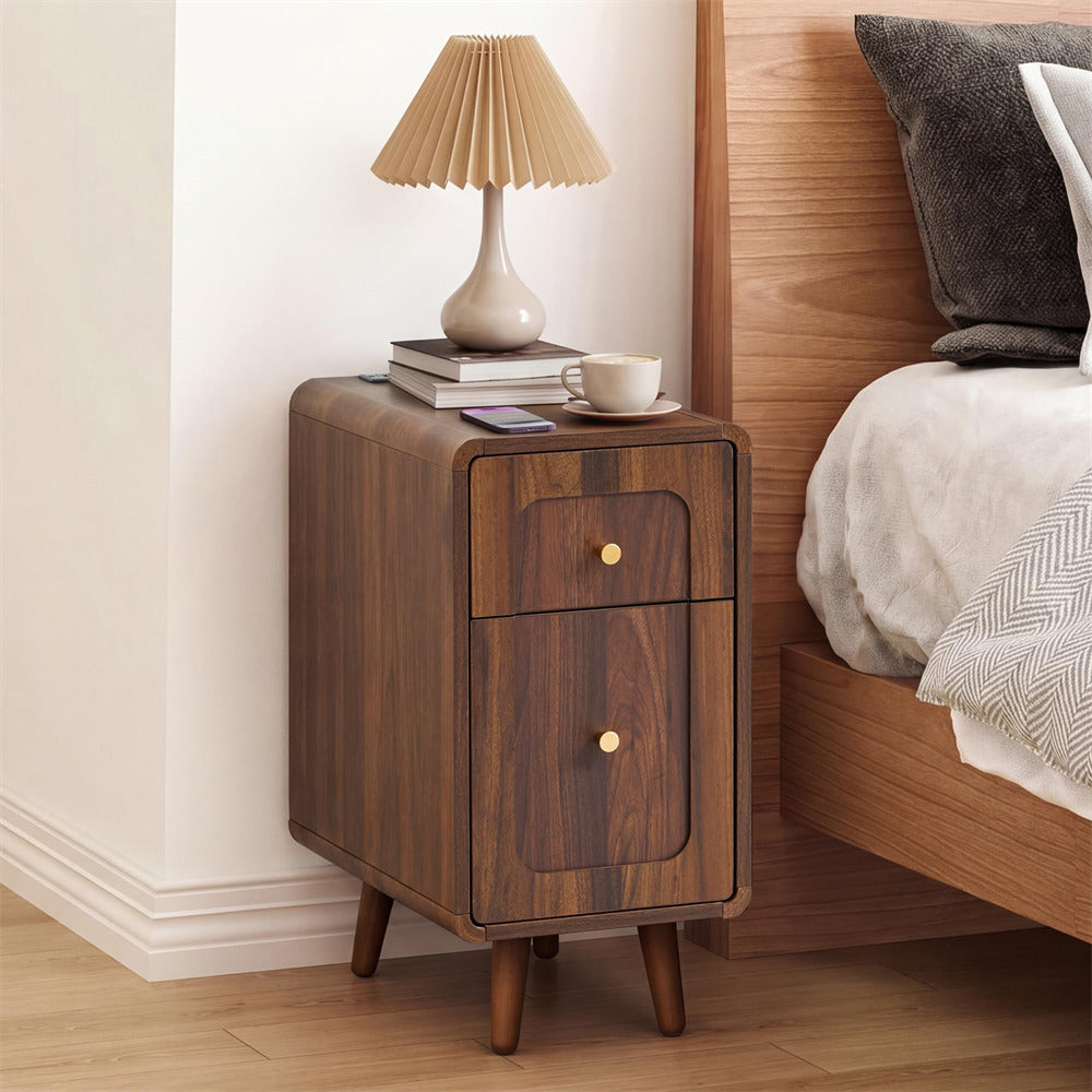 A mid-century modern wooden nightstand with two drawers and gold knobs, styled next to a bed with a pleated table lamp on top.