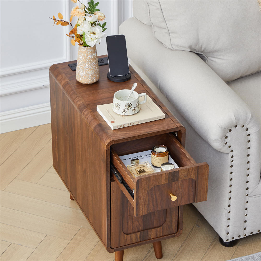 A brown wooden side table with an open drawer and a top surface, styled next to a light gray sofa on a light wood floor. The table is decorated with a vase of flowers, a smartphone, a mug, and small accessories.